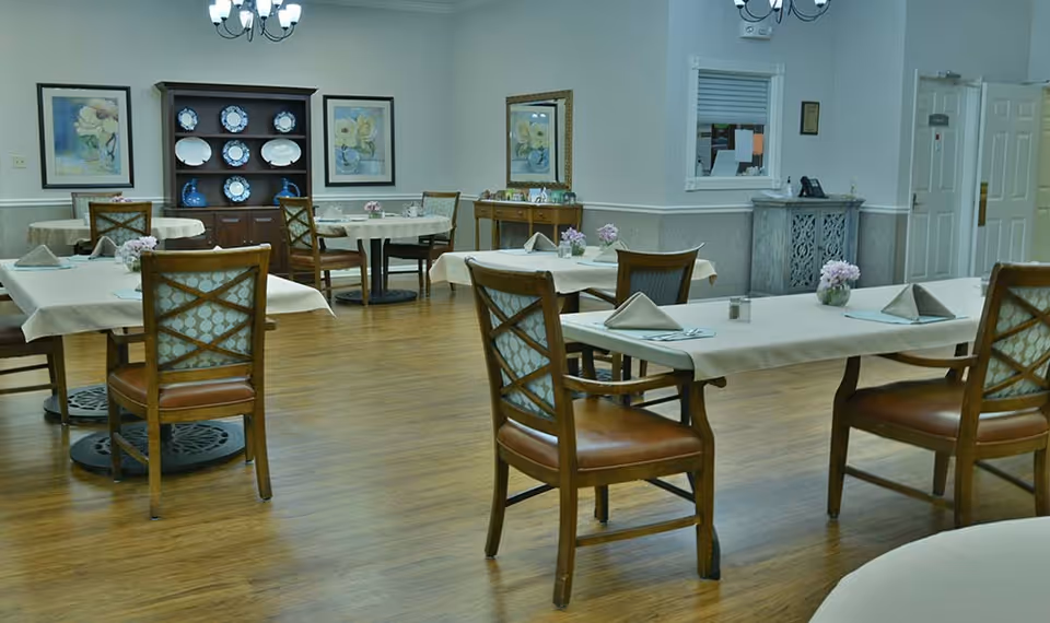 A dining room with several tables covered in white tablecloths, each set with folded napkins and small flower arrangements. Wooden chairs with patterned cushions surround the tables. The room has wooden flooring, framed floral artwork on the walls, a wooden cabinet displaying plates, and a small console table with picture frames. A chandelier hangs from the ceiling.