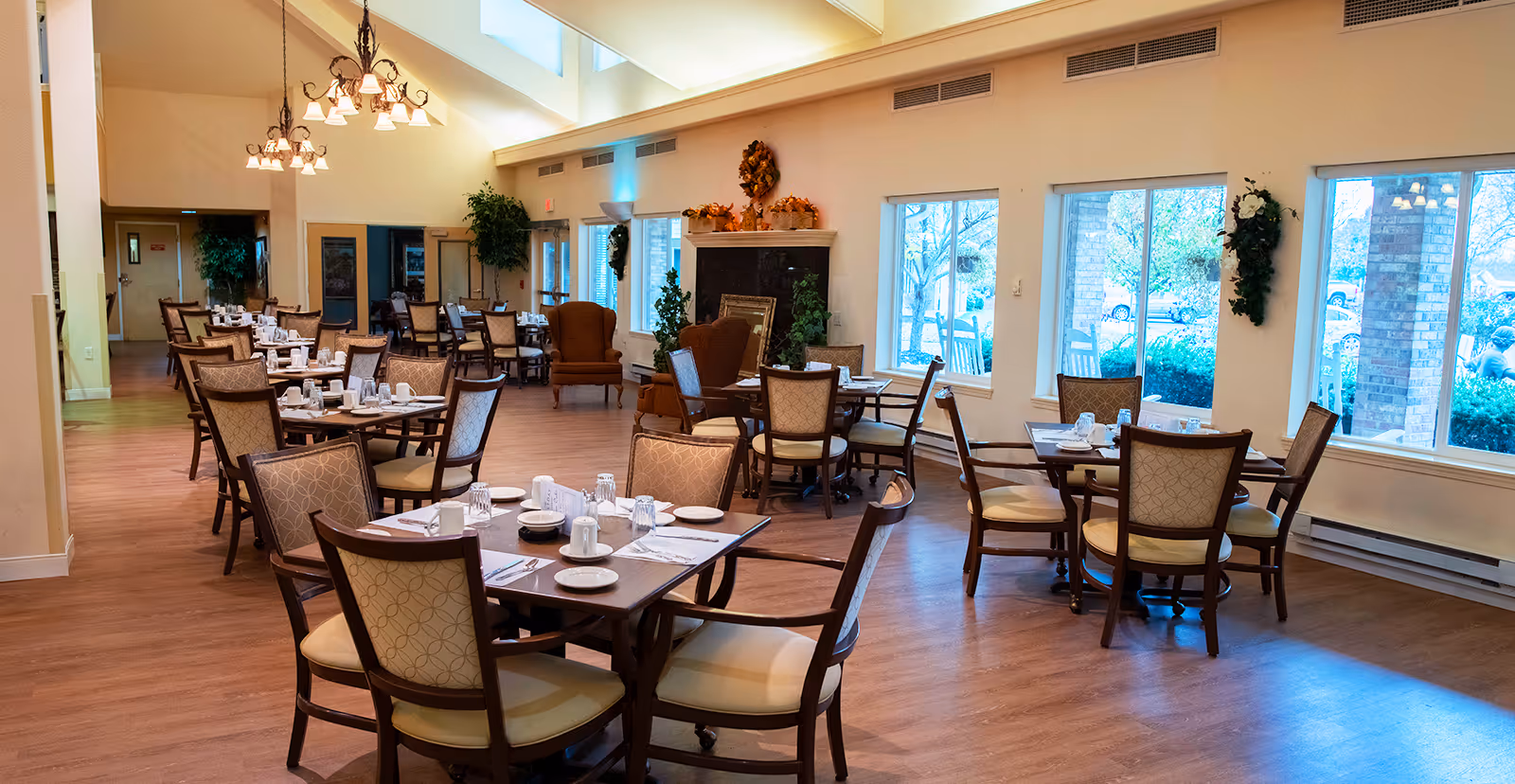 A spacious dining room with multiple tables and chairs arranged neatly. Each table is set with plates, cups, and utensils. The room features large windows letting in natural light, a fireplace decorated with autumn-themed ornaments, and chandeliers hanging from a high ceiling.