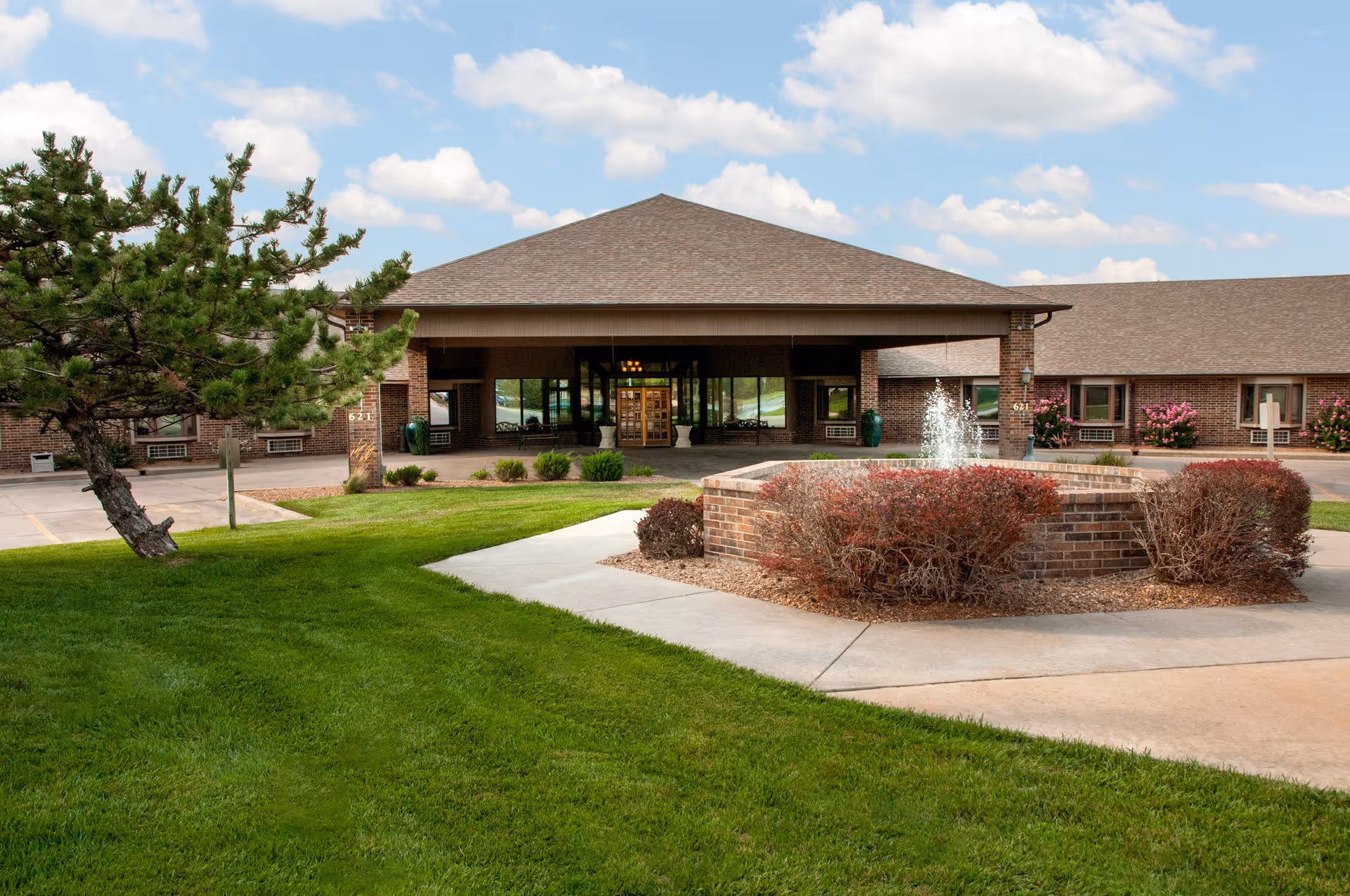 Front exterior view of Life Care Center of Andover showing a brick building with a covered entrance, a circular fountain surrounded by bushes, a well-maintained lawn, and a partly cloudy sky.