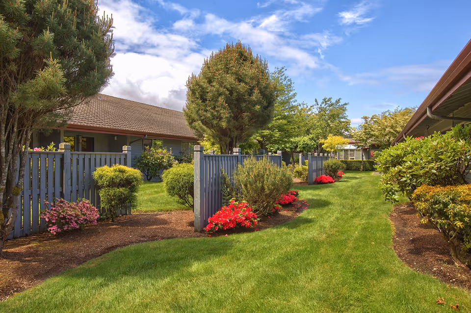 Landscaped courtyard with a green lawn, flower beds, shrubs and a wooden fence between single-story buildings under a blue sky.