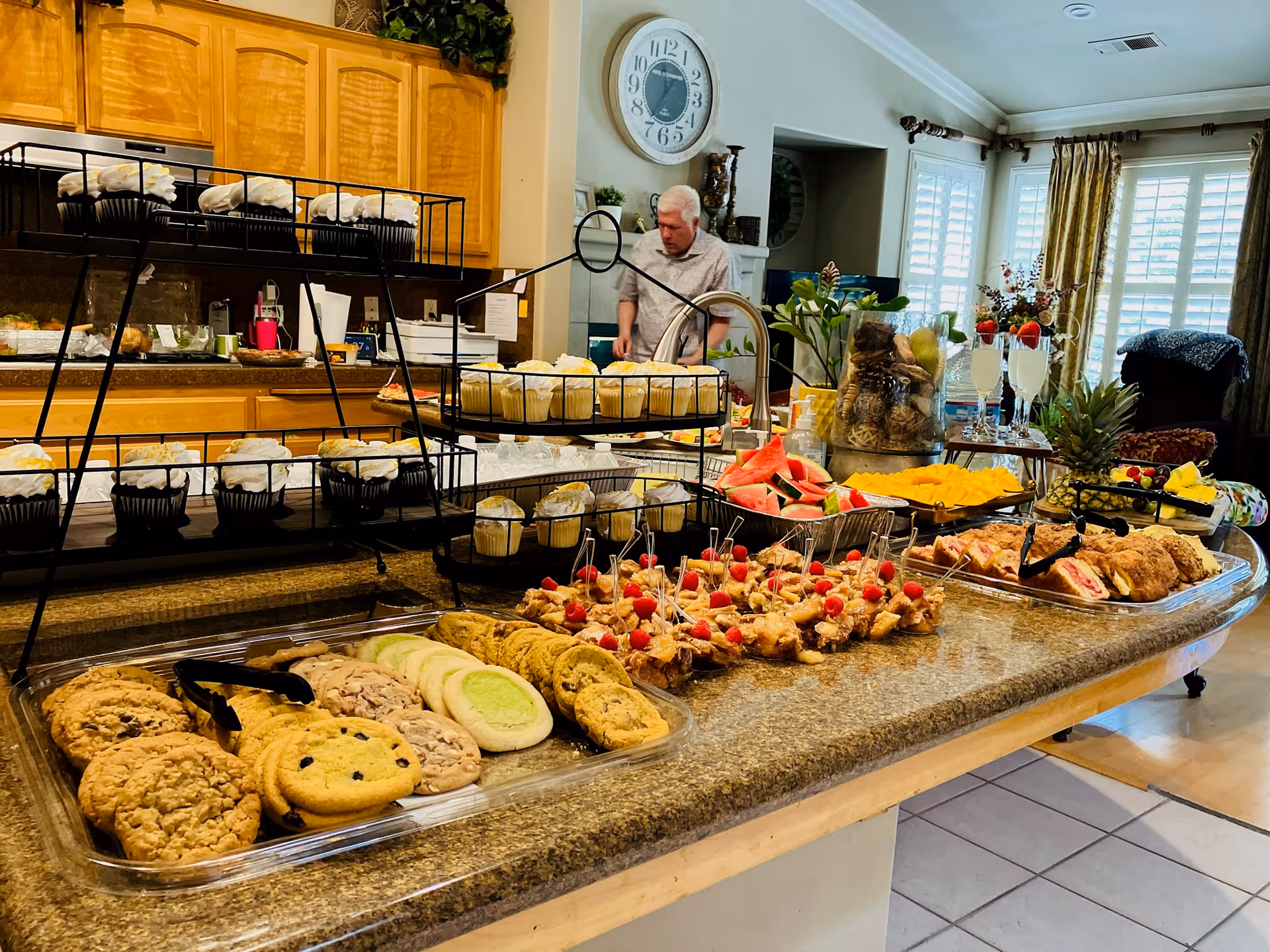 A kitchen counter filled with a variety of food items including cookies, cupcakes, fruit slices, and appetizers with toothpicks. In the background, a man is standing near the kitchen sink, and the room has wooden cabinets, a large wall clock, and windows with curtains letting in natural light.