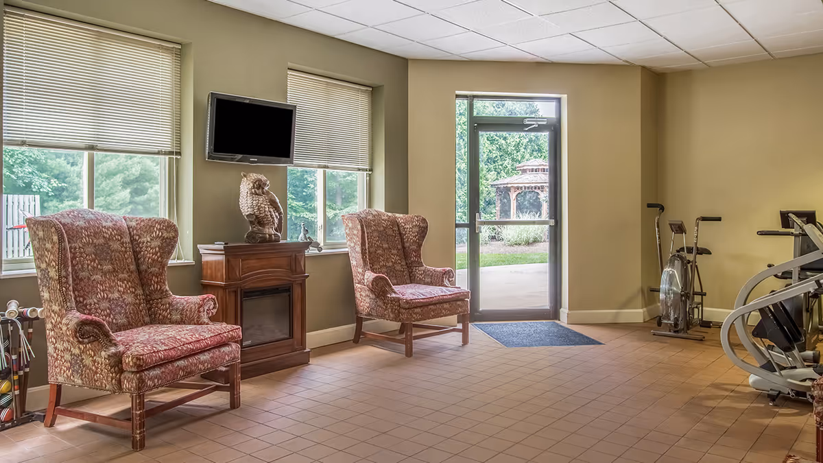 A senior living community room with two patterned armchairs, a small wooden electric fireplace with an owl statue on top, a wall-mounted TV, and exercise equipment. There are windows with blinds and a glass door leading outside to a garden with a gazebo.