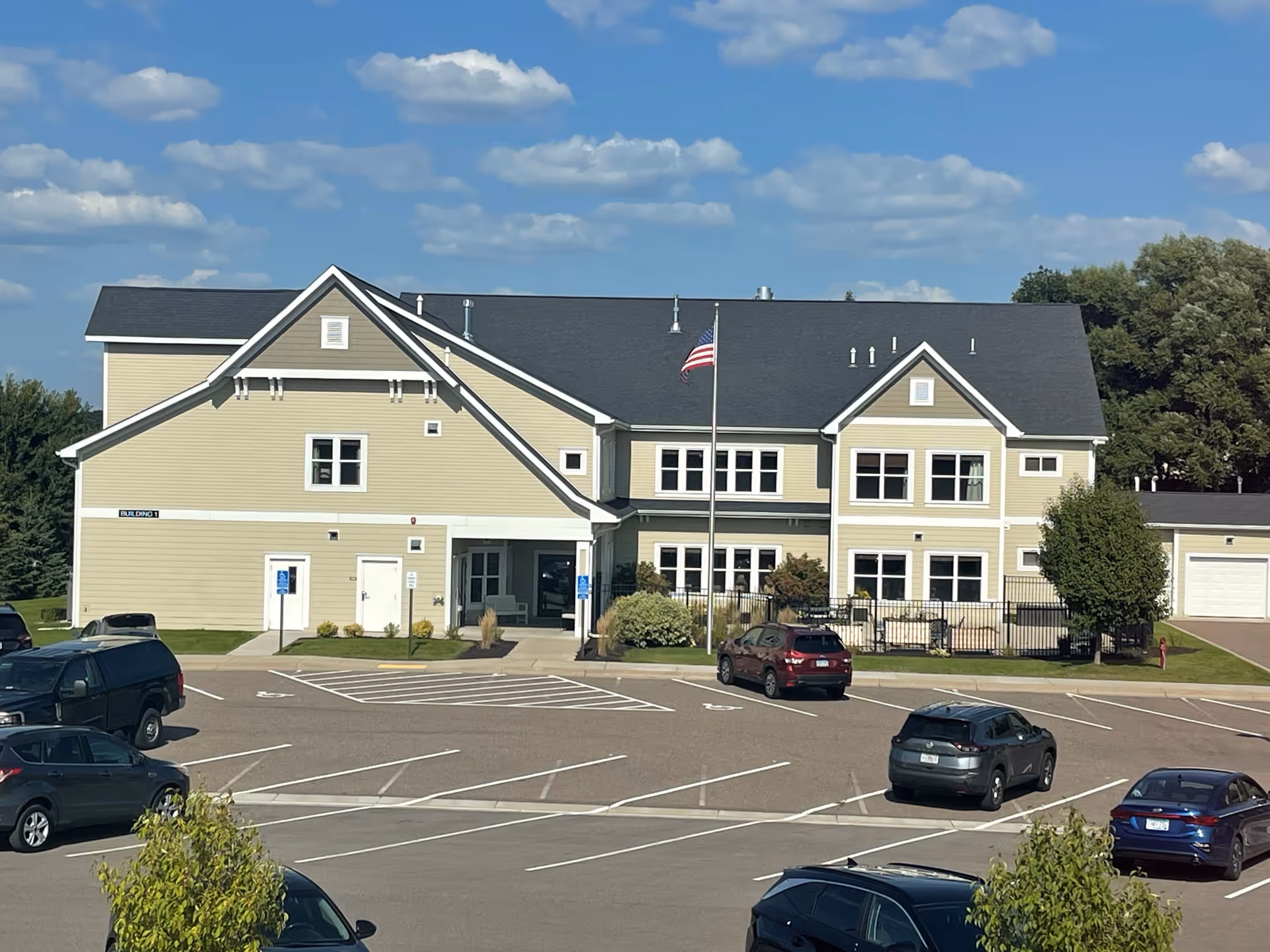 Two-story beige senior living building with an American flag and a mostly empty parking lot under a blue sky.