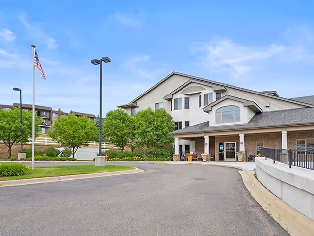 Exterior view of a senior living facility building with a driveway, green trees, an American flag on a flagpole, and a clear blue sky.
