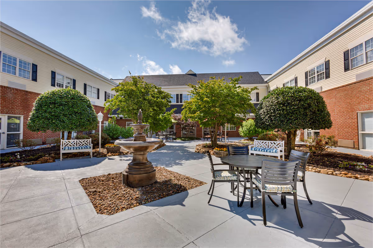Sunny courtyard with a central fountain, patio table and chairs, benches, and trimmed trees surrounded by a two-story brick-and-siding building.