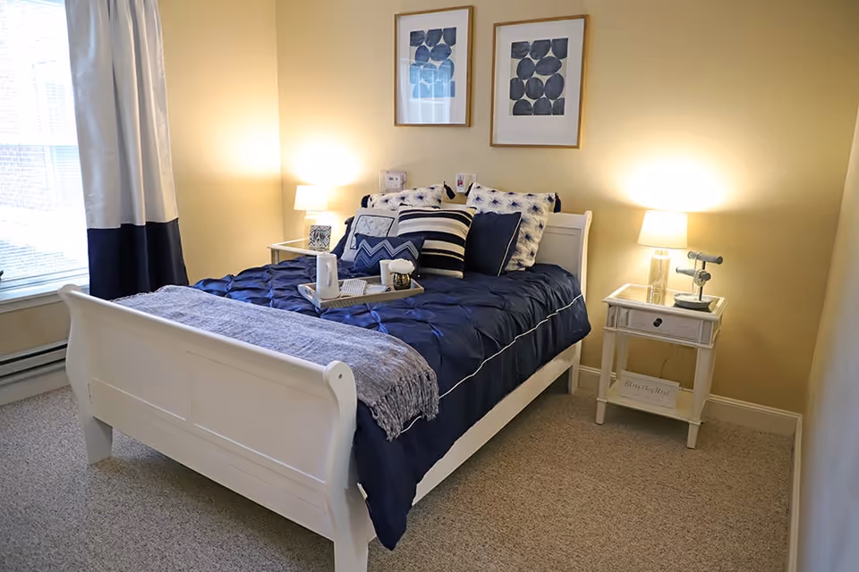 A cozy bedroom with a white wooden bed frame dressed in navy blue bedding and multiple decorative pillows. There is a gray throw blanket at the foot of the bed and a tray with a teapot and cup on the bed. Two nightstands flank the bed, each with a lamp and decorative items. The walls are painted light yellow, and two framed abstract art pieces hang above the bed. A window with white and navy curtains is on the left side of the room.