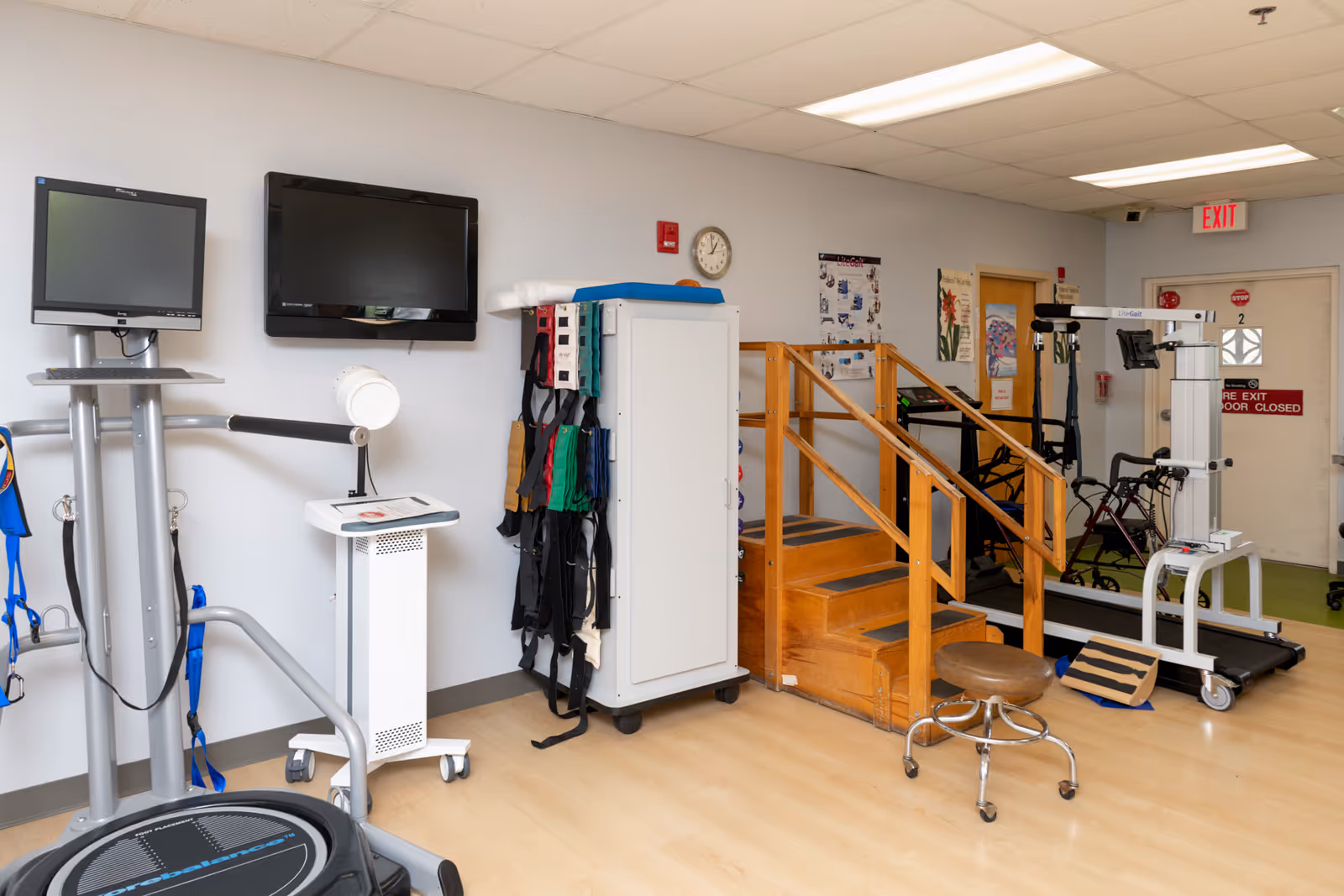 Physical therapy room with exercise equipment including a treadmill, balance platform, wooden stairs with handrails, and various therapy tools. Two monitors are mounted on the wall, and a clock is visible above a cabinet. The room has light-colored walls and flooring, with an exit door in the background.