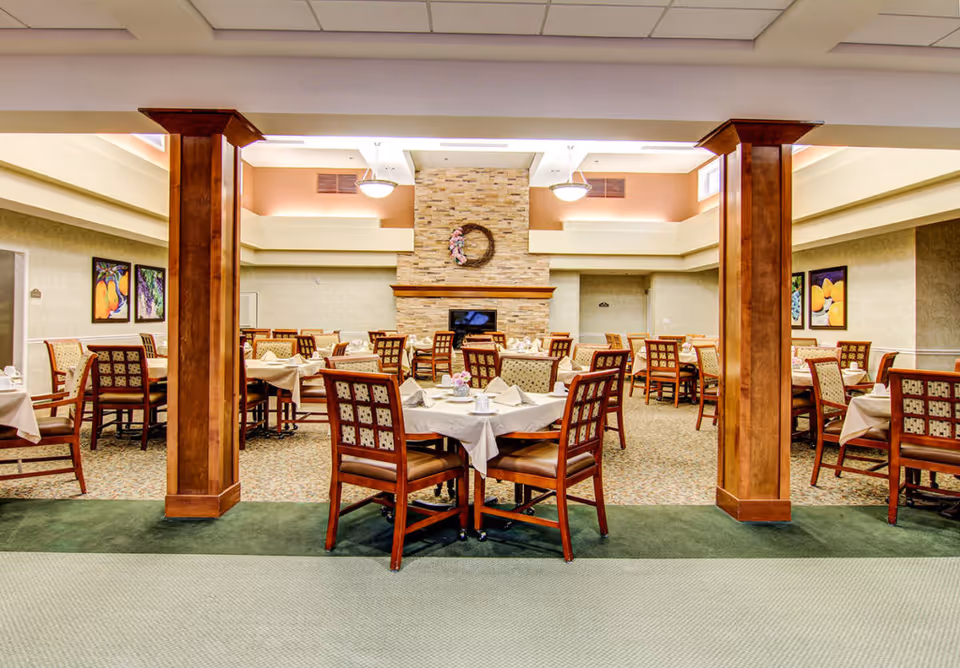 A spacious dining room with multiple tables covered in white tablecloths and set with napkins and cups. The room features wooden chairs with patterned upholstery, a stone fireplace with a decorative wreath above it, and warm lighting from ceiling fixtures. The walls are adorned with colorful framed artwork.