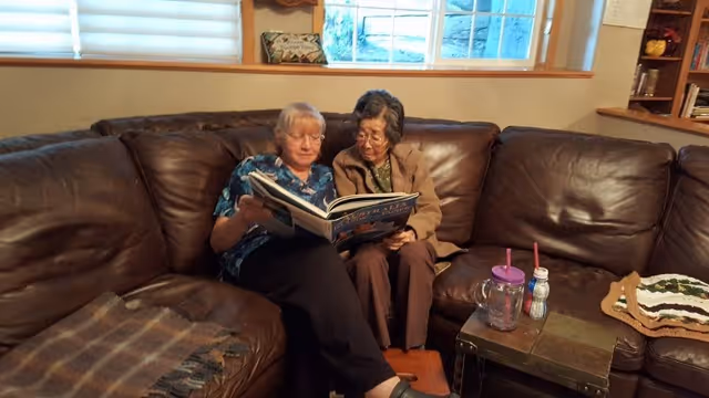 Two elderly women sitting closely together on a brown leather sectional sofa in a living room, looking at a large open book. The room has a window with blinds and a wooden shelf with decorative items. A small table in front of them holds two drinking cups and a crocheted doily.