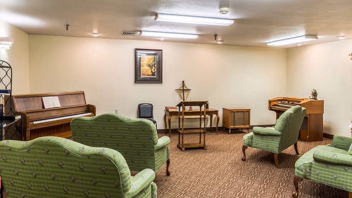 A cozy music room with vintage green upholstered chairs and sofas arranged facing a wooden piano and an organ. There is a small wooden lectern and a framed picture on the beige wall. The room has carpeted flooring and fluorescent ceiling lights.