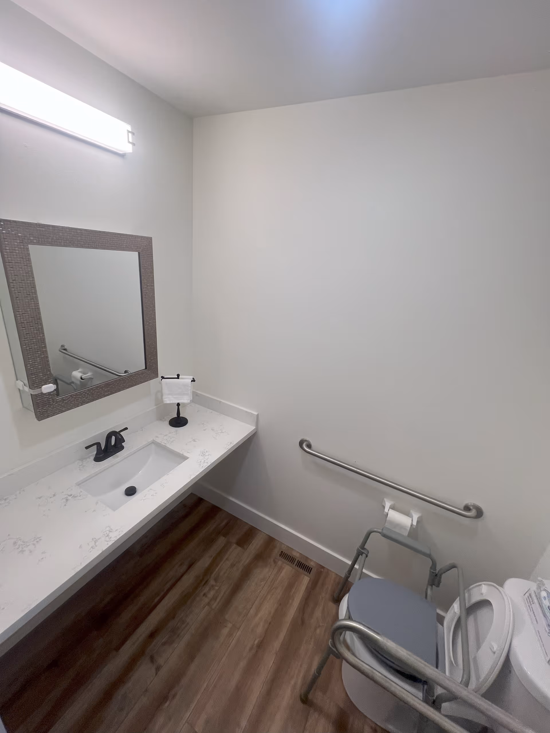 A clean, accessible bathroom with a white countertop and sink, a large mirror with a textured frame, a grab bar on the wall, a toilet with a raised seat and support rails, and wood-look flooring.
