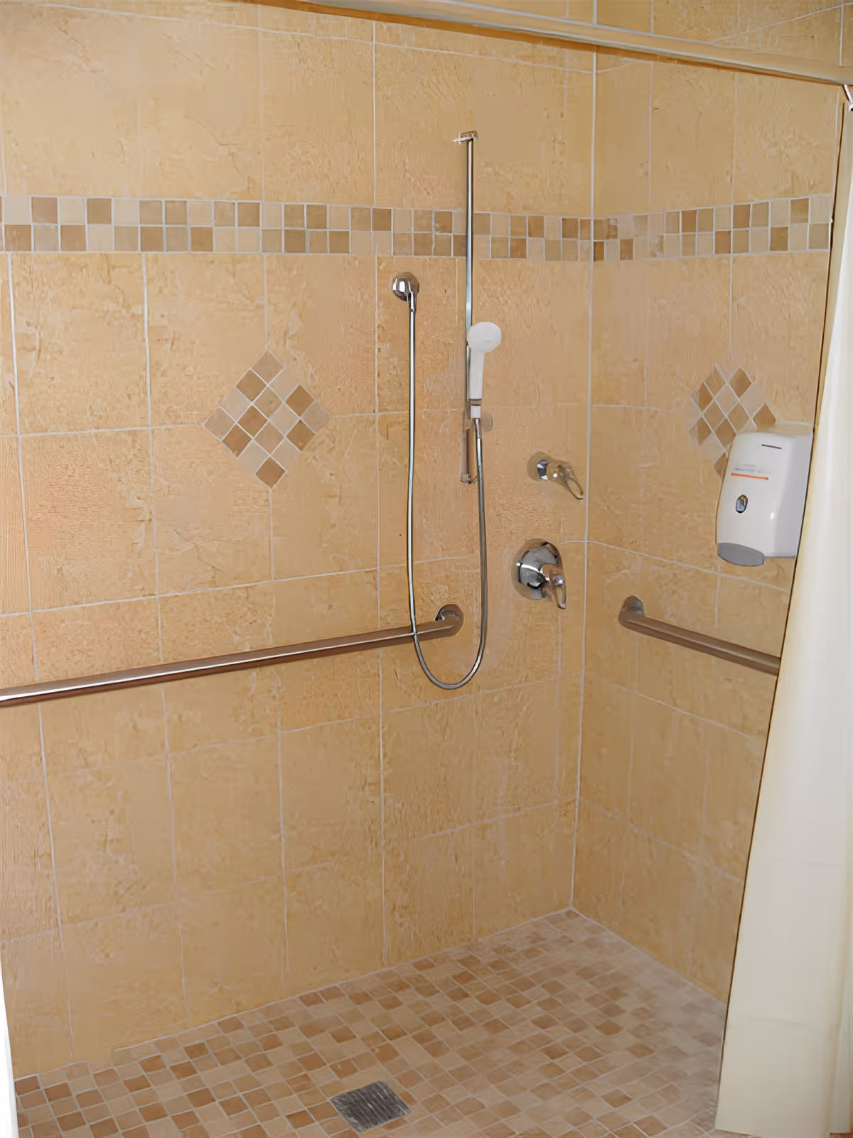 A tiled shower area with beige and brown square tiles, featuring a handheld showerhead attached to a vertical metal bar, two horizontal grab bars on the walls, a soap dispenser mounted on the wall, and a beige shower curtain partially visible on the right side.
