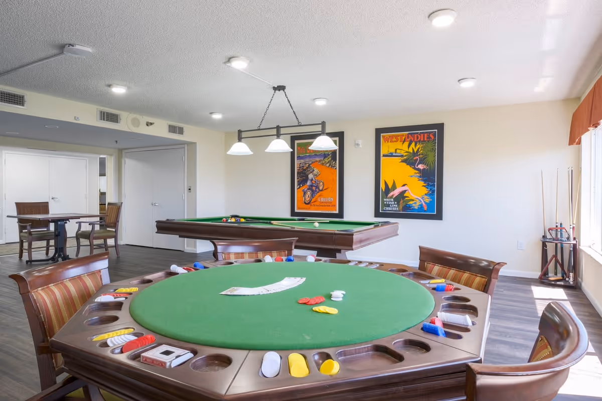 A bright game room featuring a round poker table with chips and cards, a pool table with billiard balls and cues, and two colorful framed posters on the wall. The room has wooden flooring, several chairs, and large windows letting in natural light.