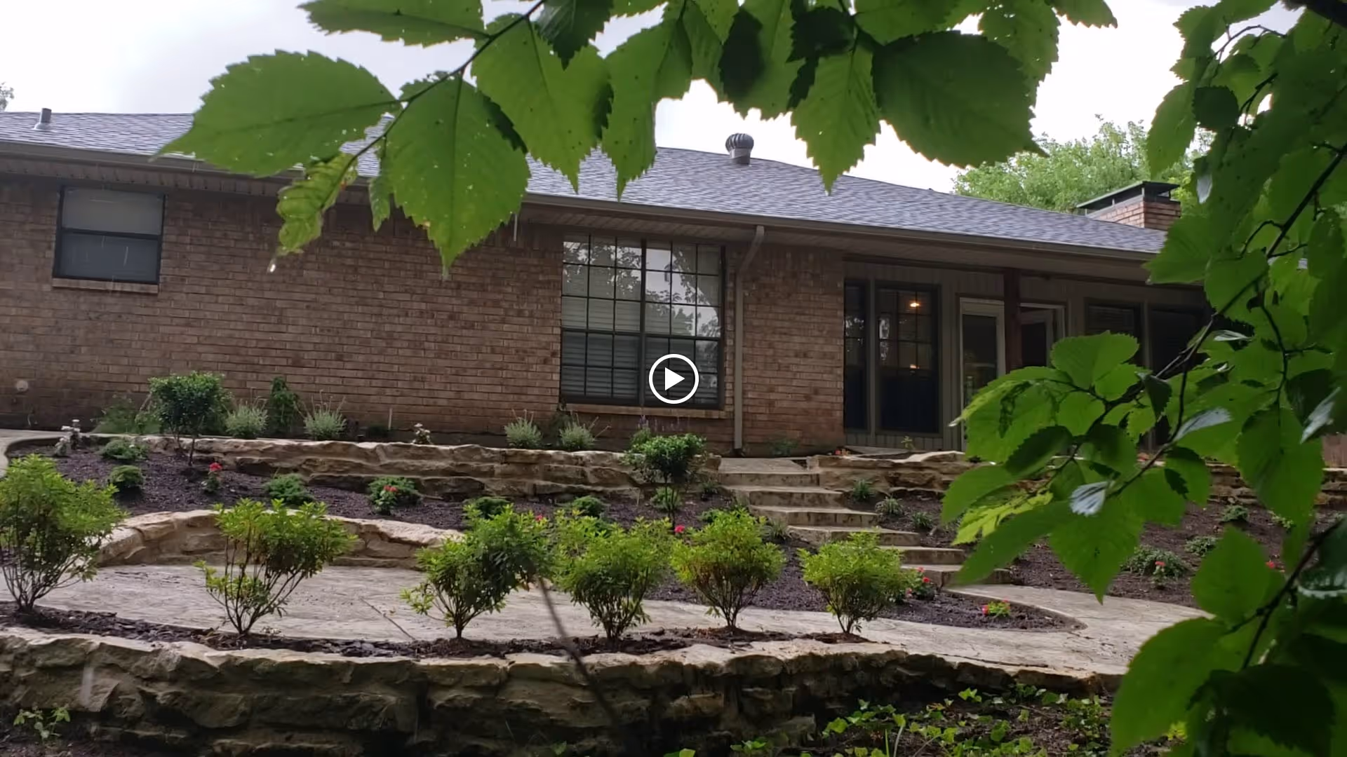 Rear exterior of a brick senior living building with terraced stone patio, steps, and landscaping framed by tree leaves.