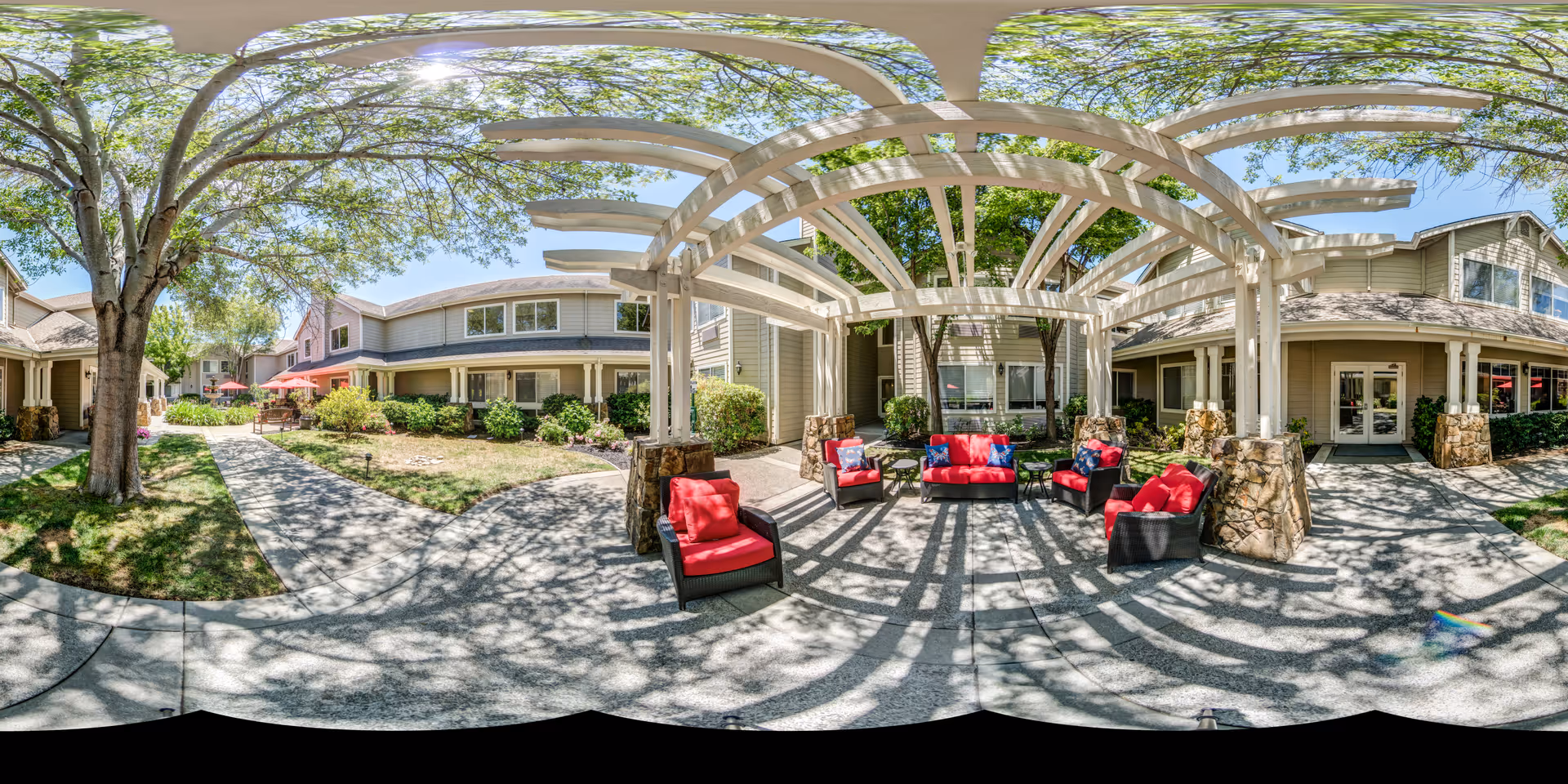 Outdoor seating area at Aegis Living Corte Madera featuring a white pergola casting shadows on the ground, with several cushioned chairs and a sofa with red cushions arranged around a small table. Surrounding the seating area are trees, shrubs, and pathways leading to residential-style buildings under a clear blue sky.