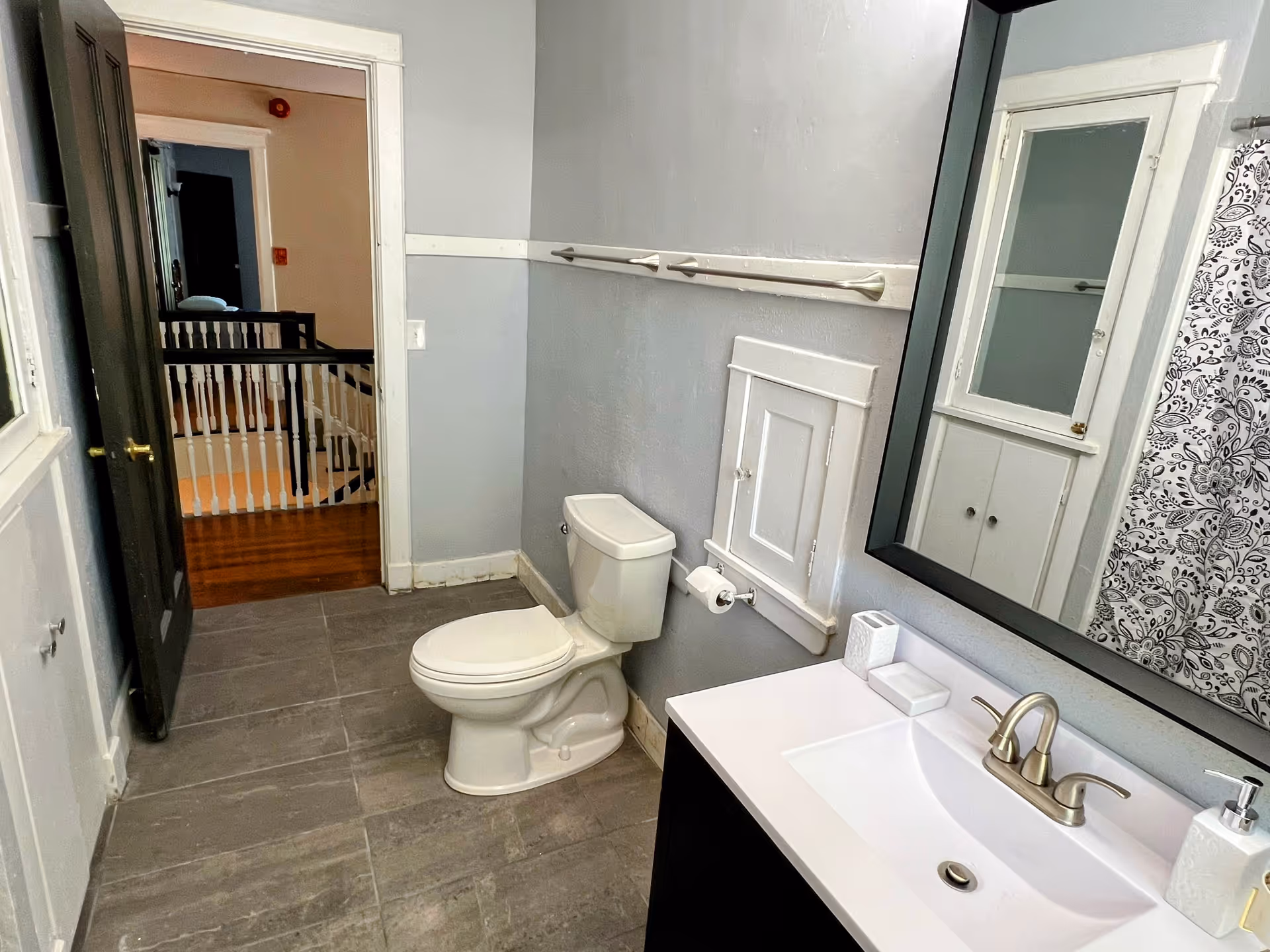 A clean bathroom with a white toilet, a white sink with a silver faucet, a large mirror with a black frame, and a white cabinet with a glass door. The walls are painted light gray, and there is a patterned shower curtain visible on the right side. The floor is tiled in gray, and the door to the bathroom is open, showing a hallway with wooden flooring and white railing.