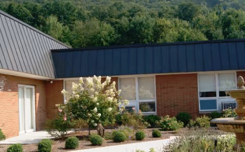 Exterior view of a single-story brick building with large windows and a metal roof, surrounded by landscaped bushes and flowering plants, with a forested hill in the background.