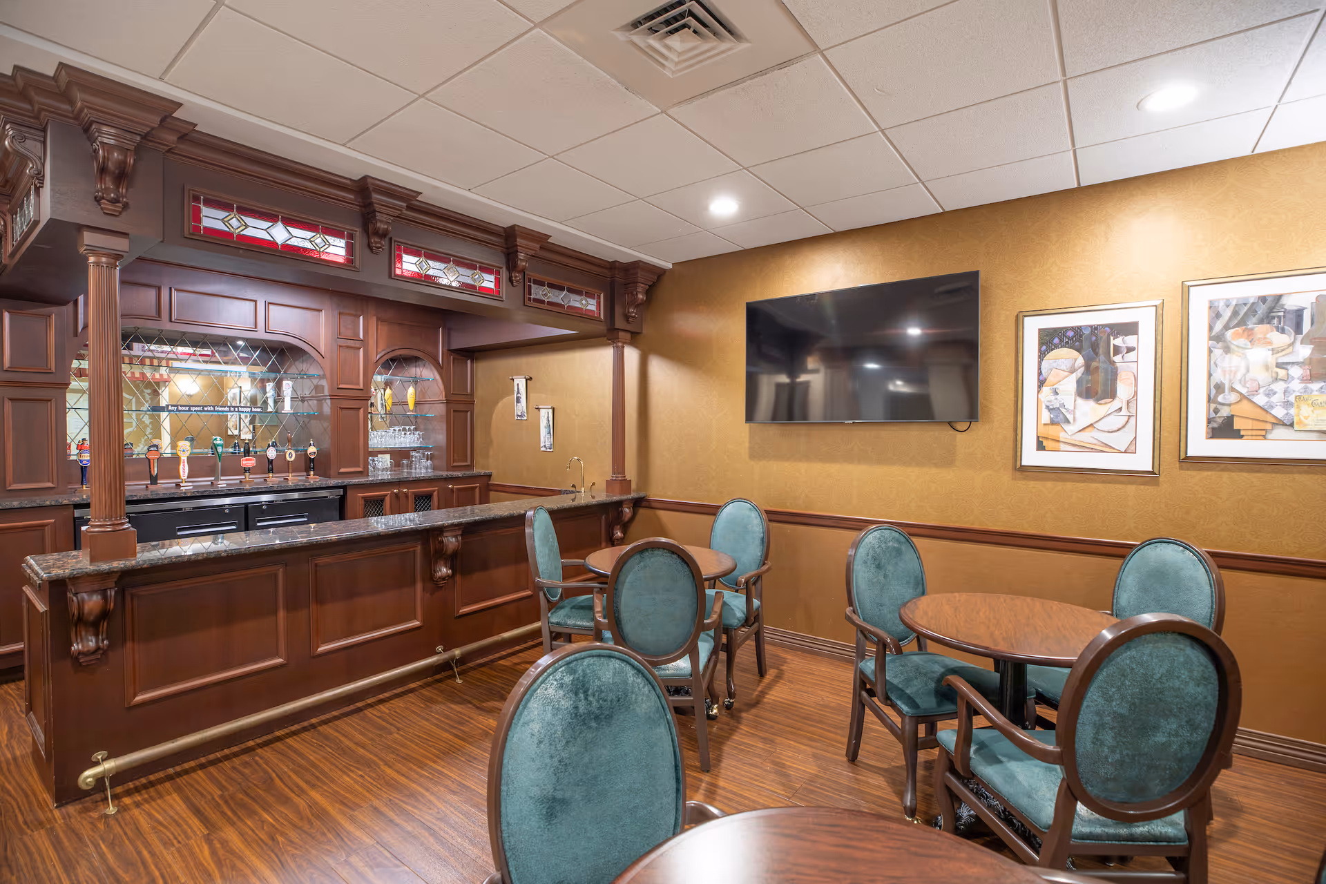 Interior view of a lounge area with a wooden bar counter featuring multiple beer taps, glass shelves with glasses, and decorative stained glass panels above. The room has several round wooden tables with green upholstered chairs, a flat-screen TV mounted on a beige patterned wall, and framed artwork hanging beside the TV.