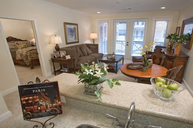 View from a kitchen counter into a living room and dining area in an apartment. The kitchen counter has a sink, a bowl of green apples, and a potted plant. The living room features a brown sofa, a side table with a lamp, and a coffee table. The dining area has a round wooden table with chairs and a decorative plant. In the background, there is a bedroom visible through an open doorway. Large windows with blinds allow natural light into the space.