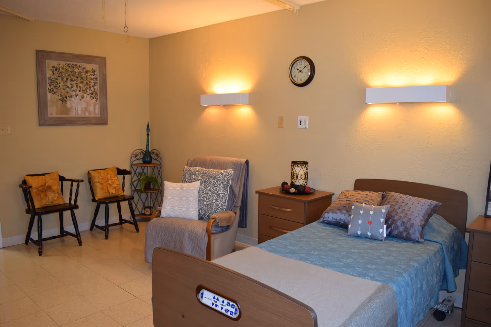 A cozy bedroom in a senior living facility with a single bed covered in blue bedding and multiple pillows. Next to the bed is a wooden nightstand with a decorative candle holder. A comfortable armchair with cushions is positioned nearby, along with two wooden chairs with orange cushions against the wall. The room has warm lighting from two wall-mounted lamps and a clock on the wall above the nightstand. A framed artwork hangs on the wall, and a small plant stand is in the corner.