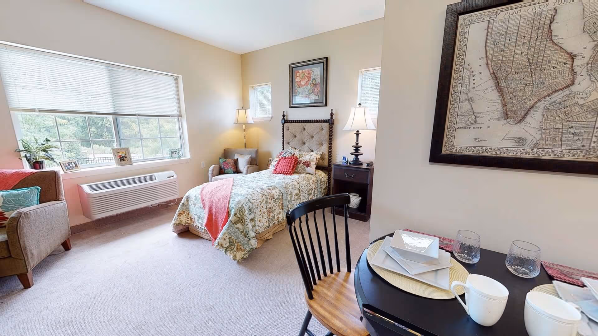 A cozy bedroom with a single bed adorned with floral bedding and a coral throw blanket. The room features a large window with blinds, a comfortable armchair with colorful cushions, two bedside lamps on either side of the bed, and a small dining table set with plates, glasses, and cups. A framed vintage map hangs on the wall near the table.