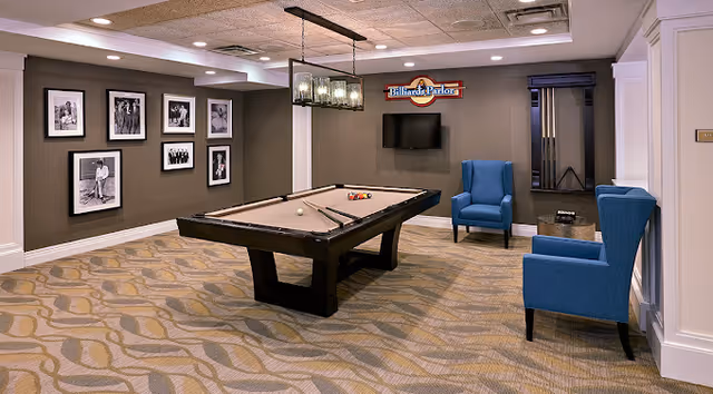 Interior billiards room with a pool table centered, two blue armchairs, a wall-mounted TV, and framed black-and-white photos.