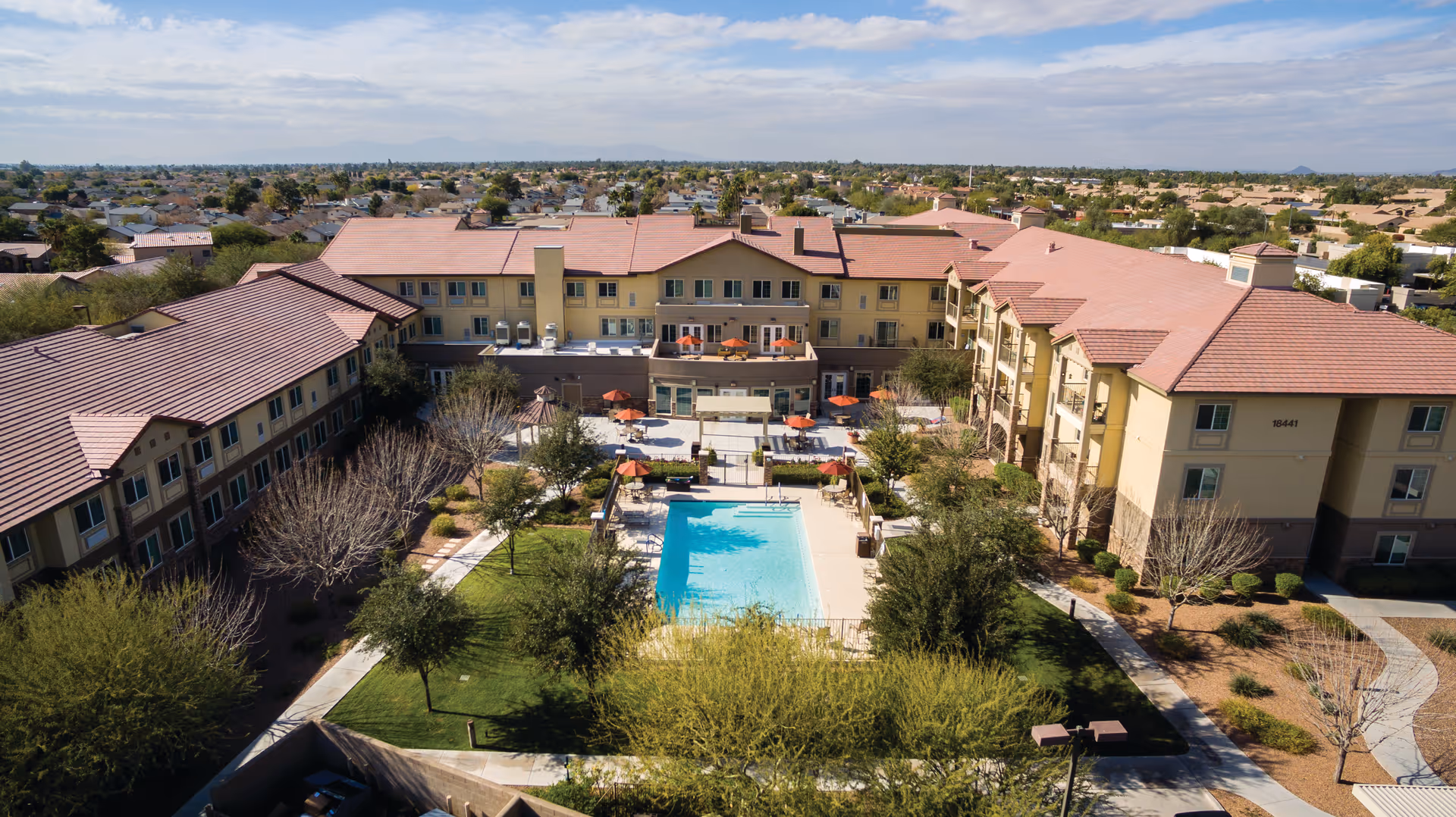 Aerial view of a senior living facility with multiple connected buildings surrounding a central courtyard featuring a swimming pool, patio seating with umbrellas, trees, and landscaped greenery under a partly cloudy sky.