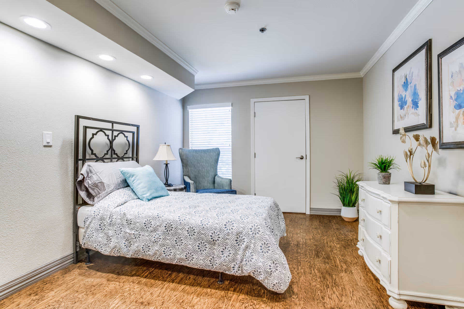 A neatly made single bed with patterned bedding and pillows in a well-lit bedroom. Next to the bed is a small table with a lamp, and a blue upholstered armchair is positioned near a window with blinds. Across from the bed is a white dresser with decorative items and two framed floral paintings on the wall. The room has wooden flooring and neutral-colored walls.