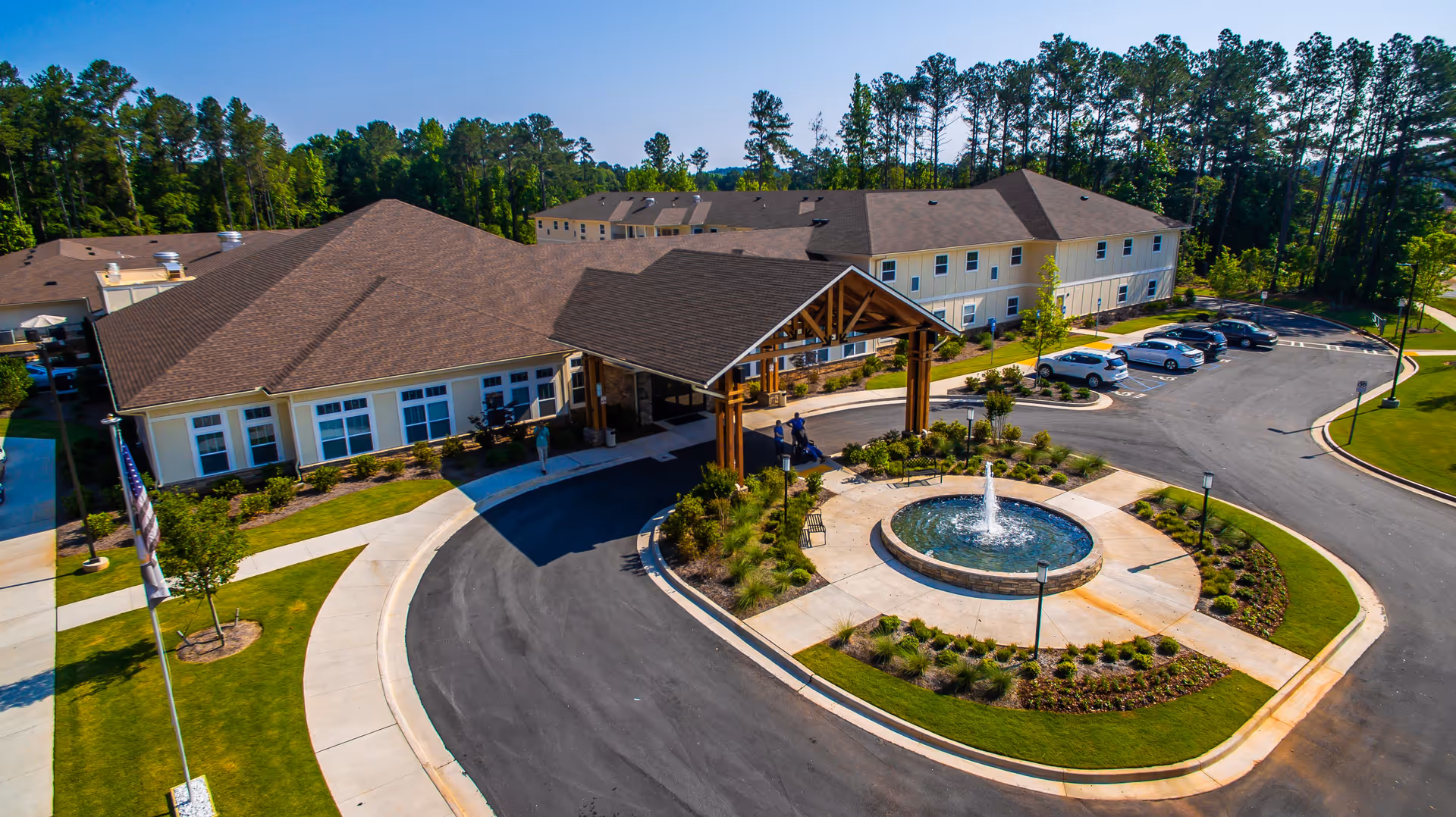 Aerial view of Oaks at Grove Park senior living facility showing the main entrance with a covered drop-off area, a circular driveway with a central fountain, landscaped greenery, and a parking lot with several cars. The building is surrounded by trees under a clear blue sky.