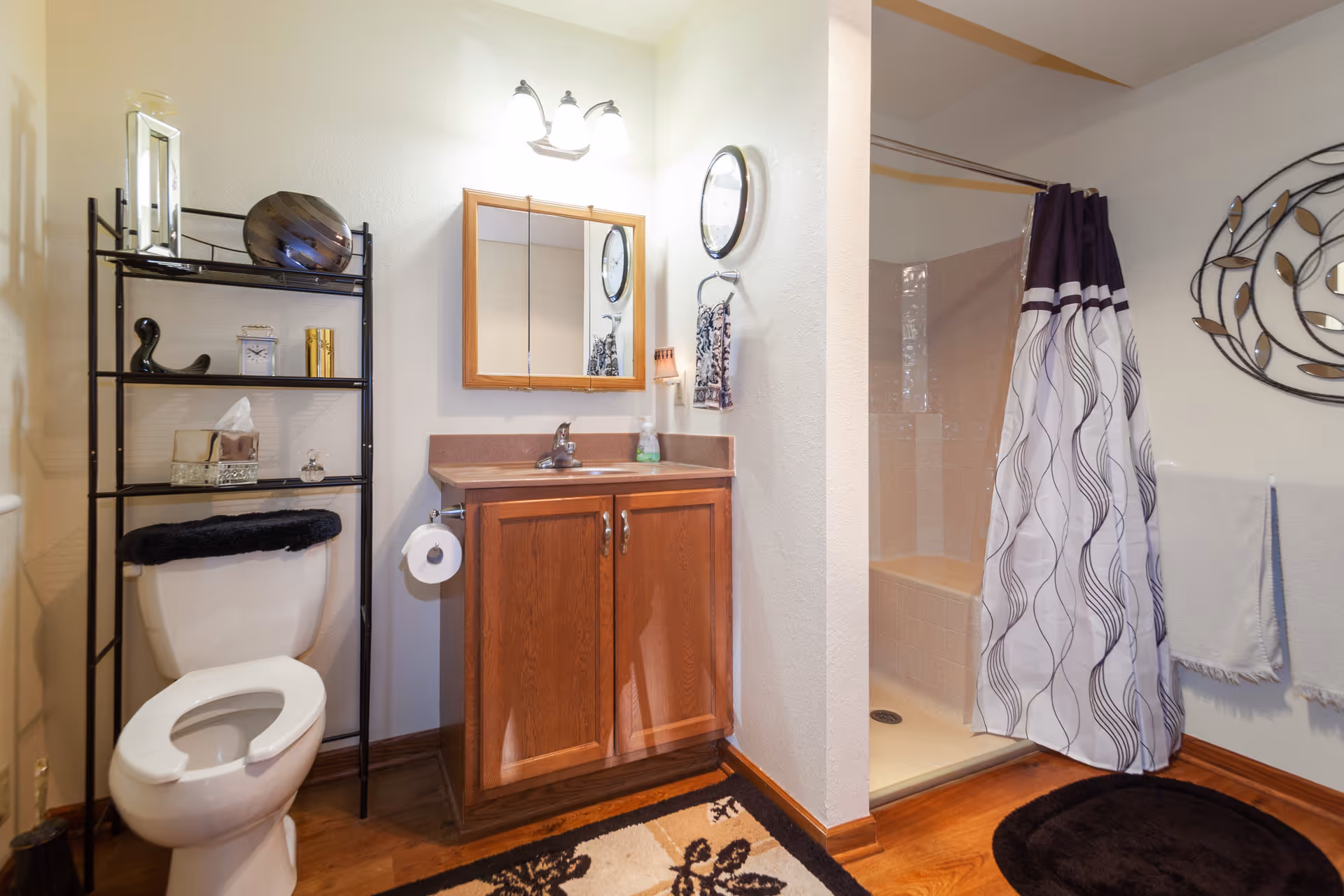 Bright bathroom featuring a toilet with shelving, a wooden vanity and mirror, and a shower with a patterned curtain.