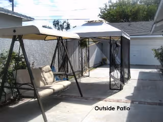 Outdoor patio area with a cushioned swing bench and a canopy tent providing shade. The patio is paved and enclosed by walls, with some plants visible along the edges. A garage door is visible in the background.