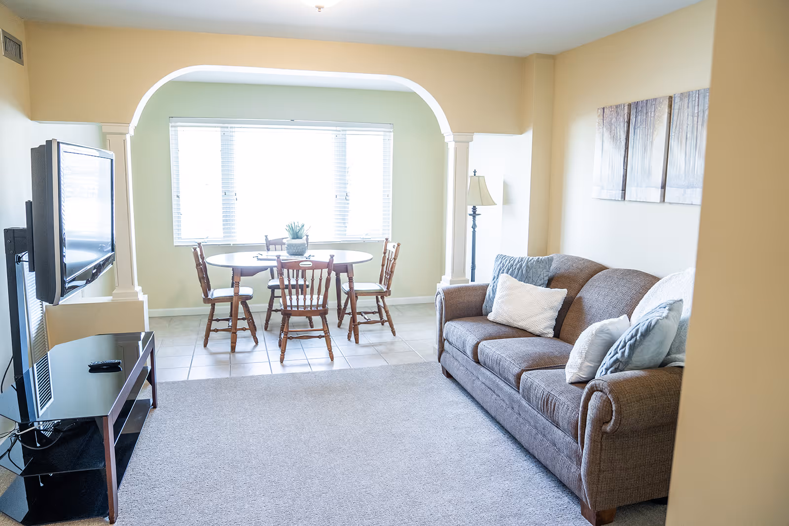 A bright living room with a brown sofa adorned with multiple cushions, a floor lamp, and a wall art piece depicting a forest. Adjacent to the living room is a dining area with a round wooden table and four matching chairs, positioned in front of a large window with white blinds. A flat-screen TV is mounted on the wall opposite the sofa, above a black glass TV stand.