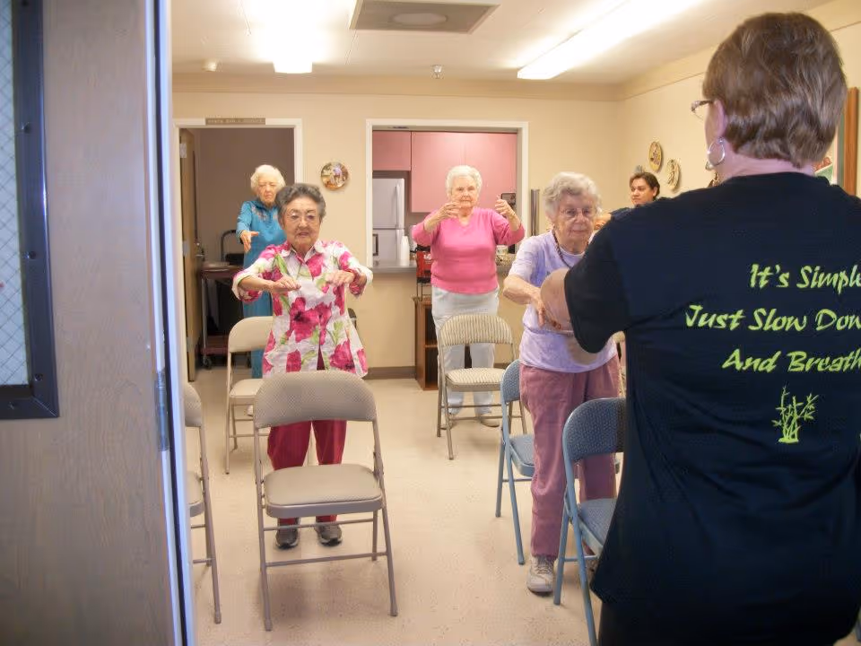 A group of elderly women participating in a seated exercise class in a room with chairs arranged in rows. An instructor wearing a black shirt with green text is leading the session. The room has beige walls and a kitchen area in the background.