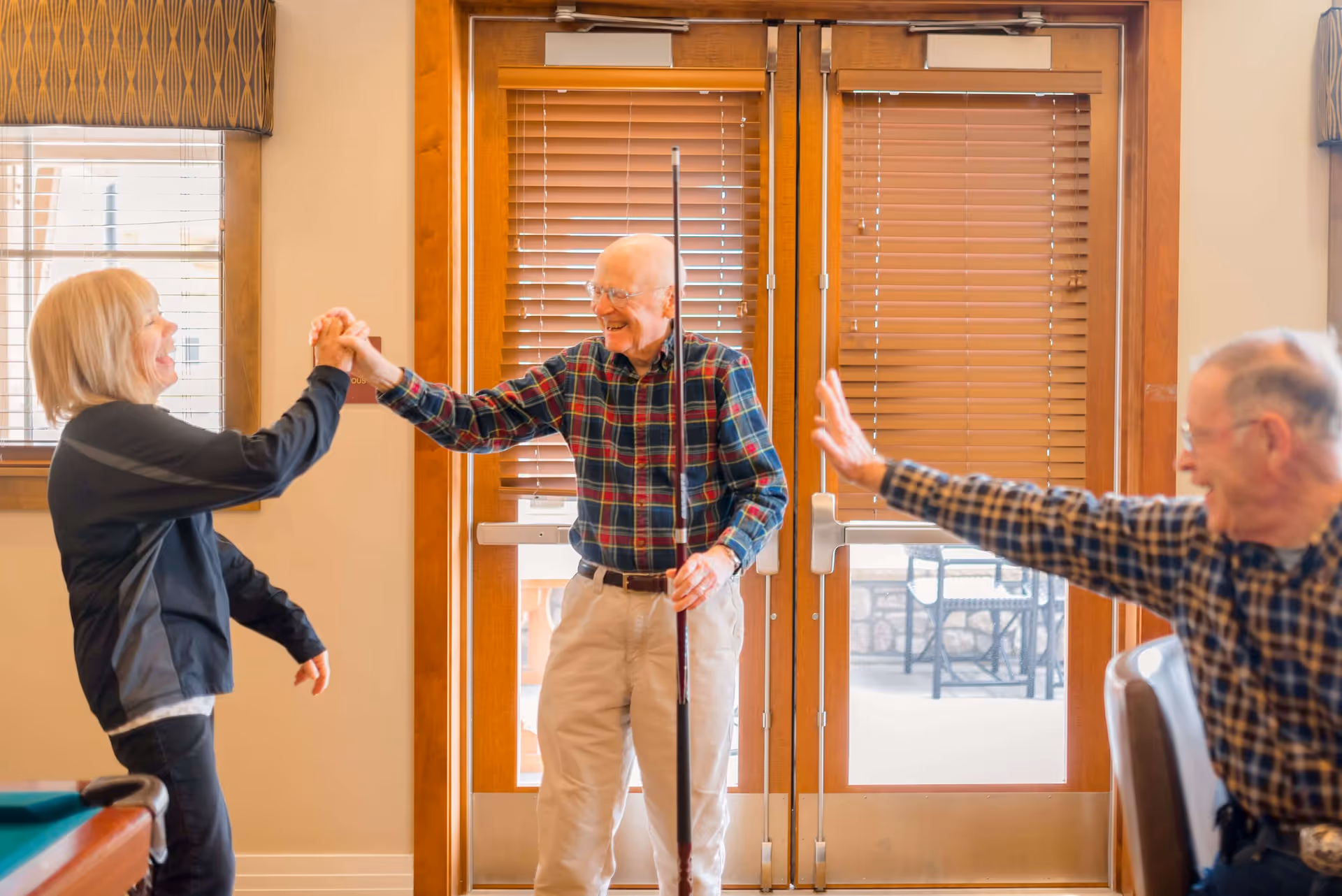 Three elderly people inside a room with wooden doors and blinds. One man in a plaid shirt is holding a pool cue and smiling while giving a high-five to a woman in a black jacket. Another man in a plaid shirt is sitting and raising his hand, appearing to wave or greet.