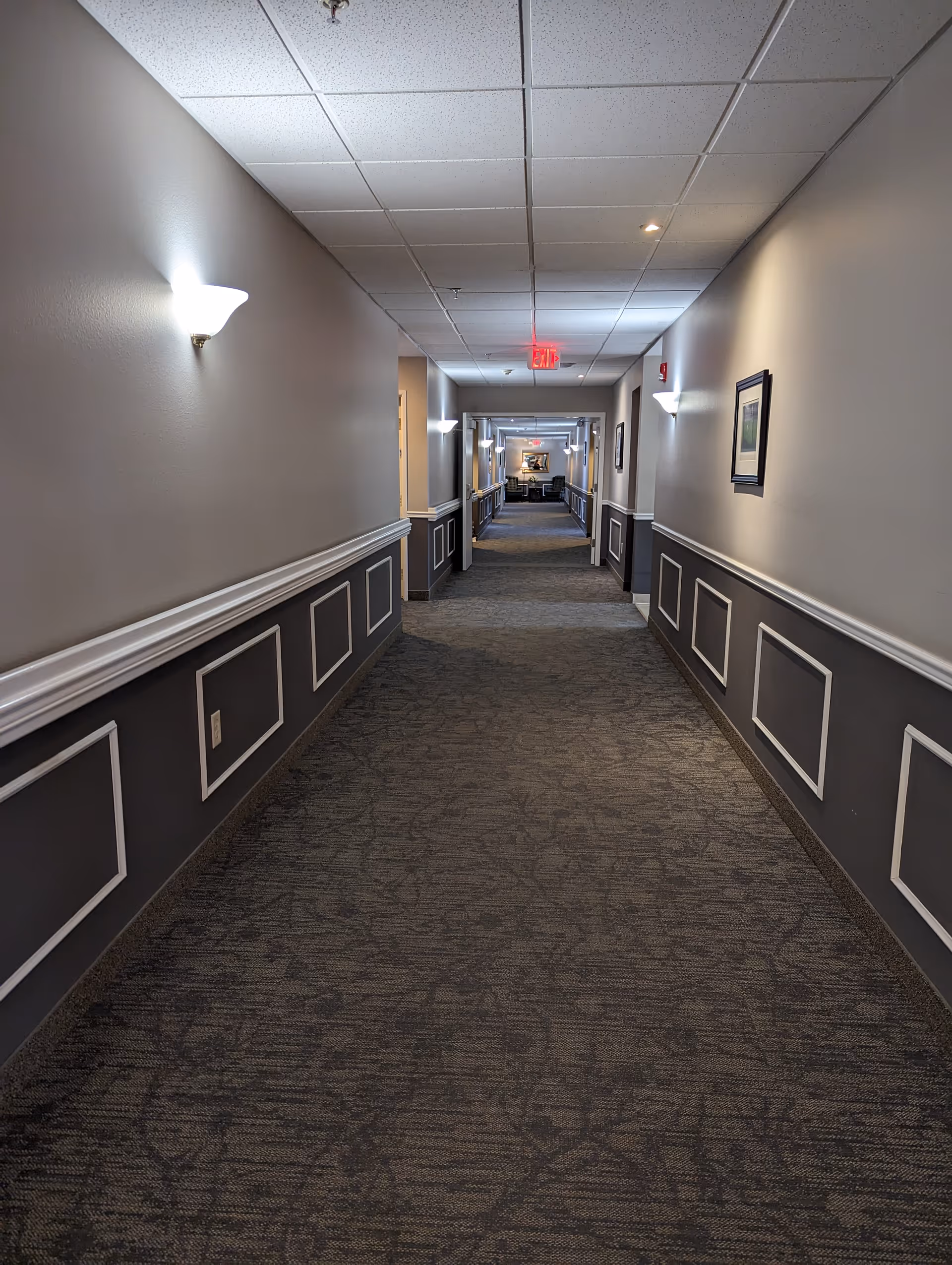A long, carpeted hallway in a senior living facility with beige and gray walls, white trim, and wall-mounted light fixtures. There are framed pictures on the walls and an exit sign at the far end of the corridor.