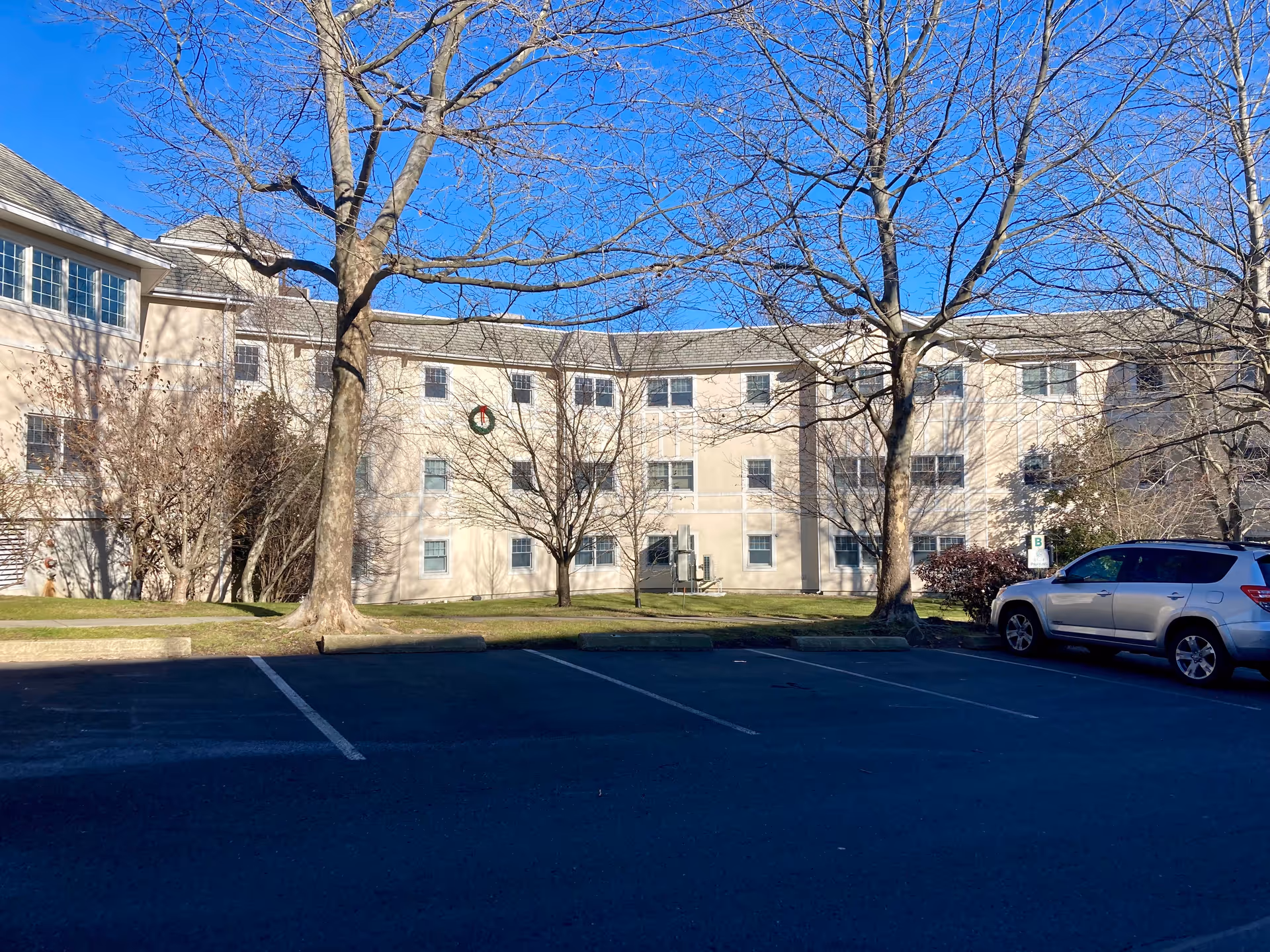 Exterior view of a three-story beige building with multiple windows, leafless trees in front, a green lawn, and a parking lot with a silver SUV parked on the right side under a clear blue sky.
