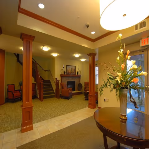 A cozy senior living facility common area with a fireplace, comfortable armchairs, wooden columns, and a round table with a floral arrangement near the entrance door.