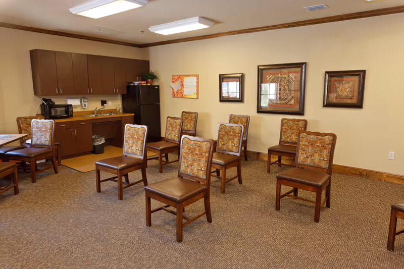 A room with multiple wooden chairs arranged in rows facing forward. The room has a kitchenette area with dark wood cabinets, a black refrigerator, a microwave, and a sink. The walls are beige with three framed decorative artworks and a bulletin board. The floor is carpeted and the ceiling has fluorescent lighting.