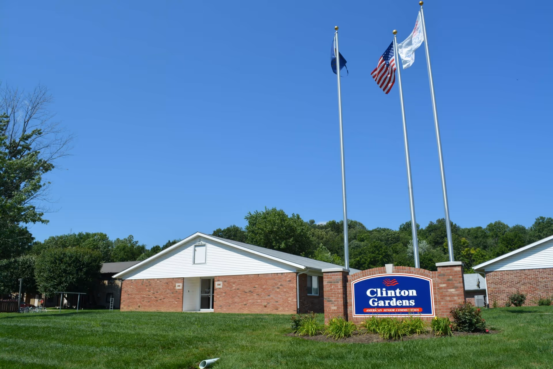 Exterior view of Clinton Gardens senior living facility with a brick building, green lawn, and three flagpoles displaying flags against a clear blue sky.