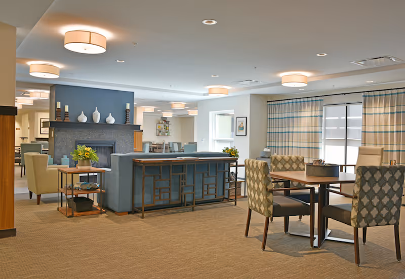 A spacious and well-lit senior living common area with a fireplace, comfortable seating including armchairs and patterned chairs around a table, decorative vases on the mantel, and large windows with striped curtains allowing natural light to enter.