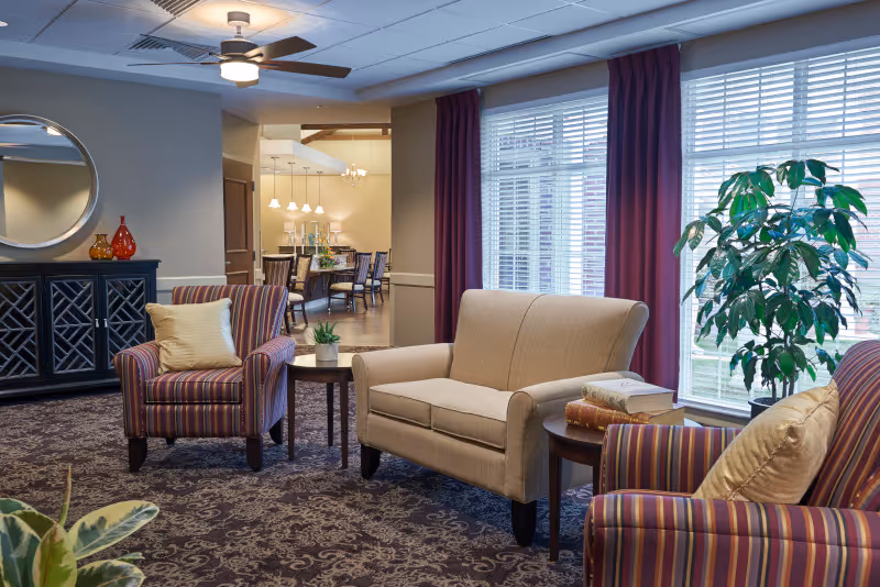 A cozy senior living community lounge area with a beige loveseat, two striped armchairs with cream pillows, a small round wooden table with books, a side table with a small plant, a large window with white blinds and purple curtains, a ceiling fan, and a dark patterned carpet. In the background, a dining area with a long table and chairs is visible.