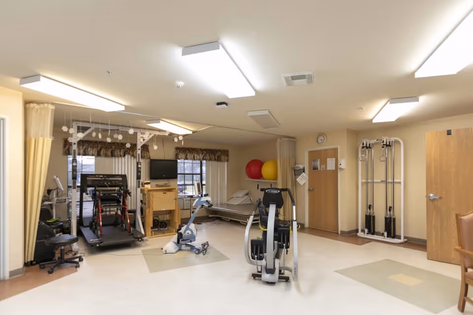 A rehabilitation room with various exercise equipment including a treadmill, stationary bike, and weight machine. There is a hospital bed with two large exercise balls behind it, a television mounted on a wooden stand, and a clock on the wall above a closed door. The room has beige walls, ceiling lights, and windows with floral valances.