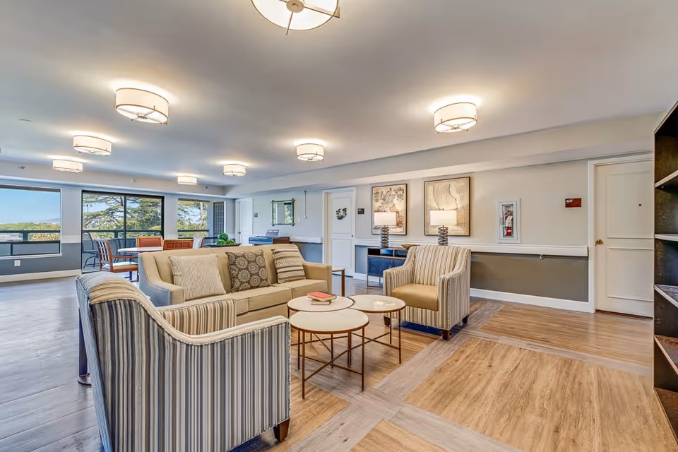 A bright and spacious senior living facility common area with a beige sofa and two striped armchairs arranged around three round nesting coffee tables. The room features wood flooring, modern ceiling lights, framed artwork on the walls, and large windows providing a view of greenery outside. There is also a dining table with chairs near the windows.