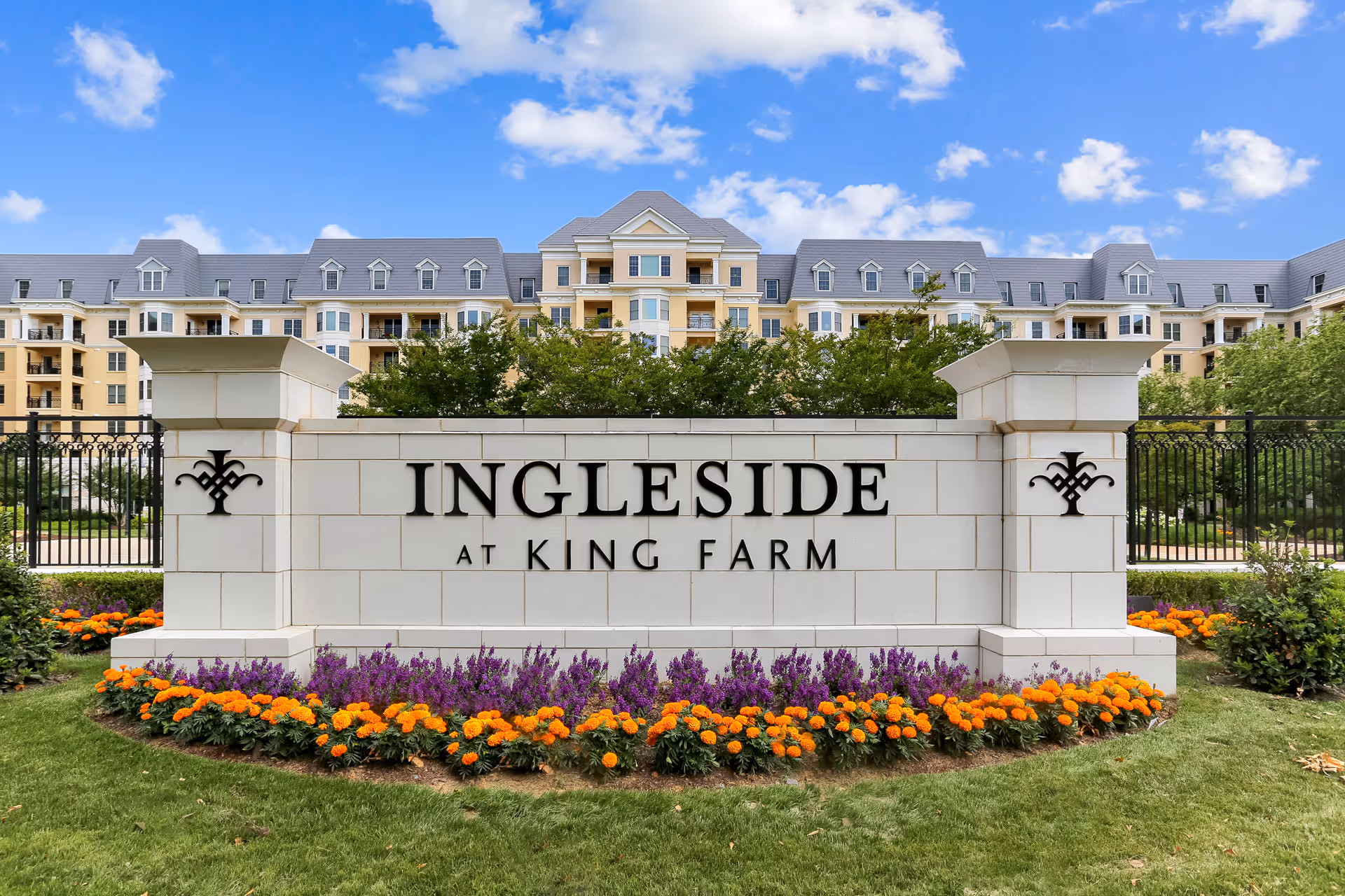 Entrance sign for Ingleside at King Farm, a senior living facility, with a large beige stone sign displaying the facility name surrounded by colorful flowers and green grass, with a multi-story residential building and blue sky with clouds in the background.