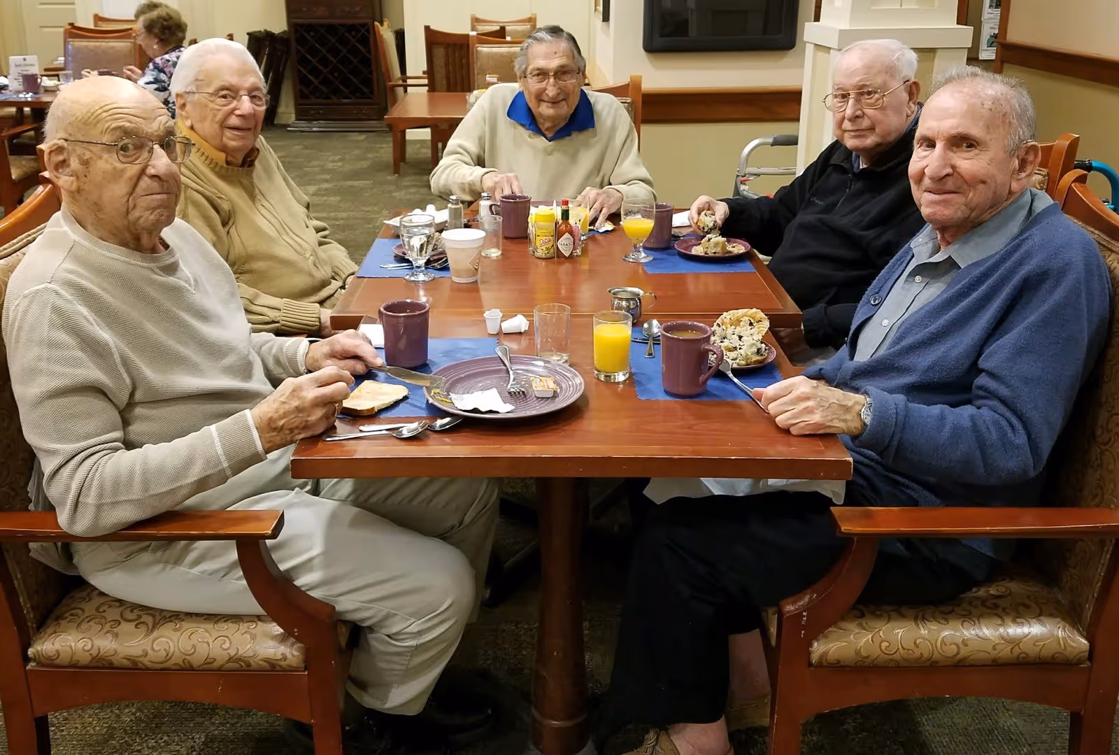 Five elderly men sitting around a dining table in a senior living facility, enjoying a meal with plates, cups, and glasses of orange juice in front of them.