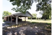 A covered carport structure with multiple parking spaces under a pitched roof, situated on a grassy area with large trees providing shade. The background shows more open green space and additional trees.
