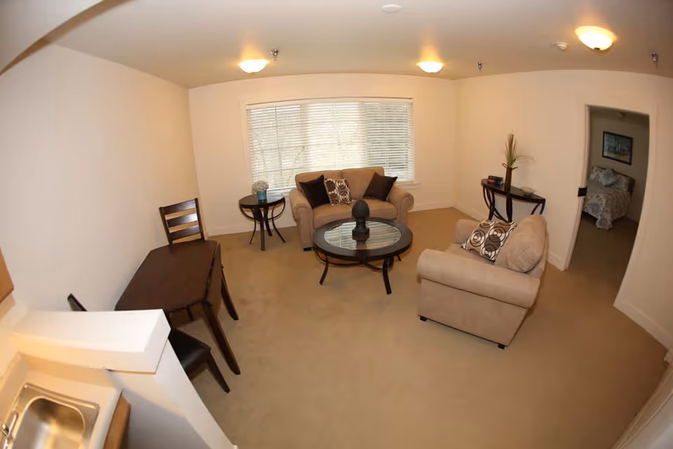 A cozy living room with beige carpet and walls, featuring a loveseat and an armchair with patterned cushions, a round glass coffee table, a small round side table with a flower vase, a wooden desk with a chair, and a console table with decorative items. A large window with blinds lets in natural light. A doorway leads to a bedroom with a bed and framed artwork on the wall.