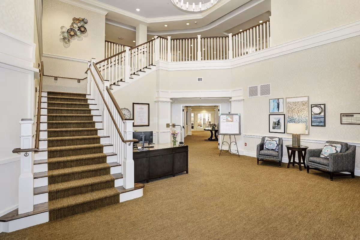 Interior view of a senior living facility lobby with a carpeted staircase leading to an upper floor. There is a reception desk with a computer and a vase of flowers, two armchairs with patterned pillows, a small side table with a lamp, and framed artwork on the walls. The area is well-lit with a chandelier and recessed lighting.