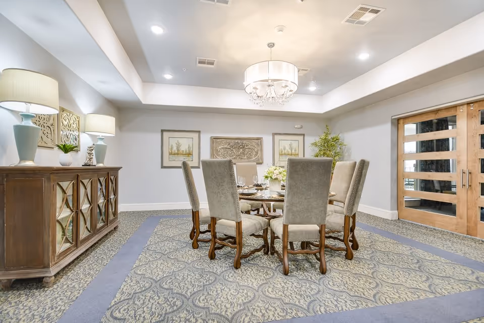A well-lit dining room with a round table set for six, surrounded by upholstered chairs. The room features a patterned carpet, a wooden sideboard with two lamps and decorative items, framed artwork on the walls, a chandelier hanging from the ceiling, and double wooden doors with glass panels.
