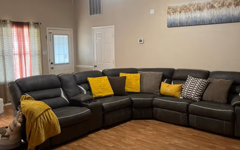 A living room with a large dark gray sectional sofa adorned with yellow, brown, and patterned throw pillows. A yellow blanket is draped over one arm of the sofa. The room has wood flooring, beige walls, a door with a window, and a window with sheer curtains. A decorative painting hangs on the wall above the sofa.